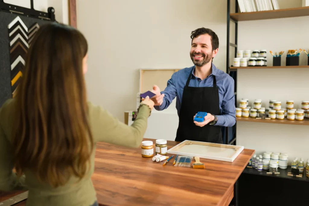 a-jovem-cliente-esta-entregando-ao-vendedor-sorridente-seu-cartao-de-credito-para-pagar-seus-suprimentos-de-arte-na-loja-pequenos negócios que dão certo com pouco dinheiro