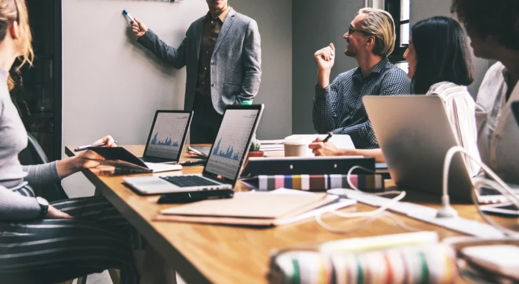 funcionários em mesa de reunião em grupo observando quadro negro - gestão ecommerce