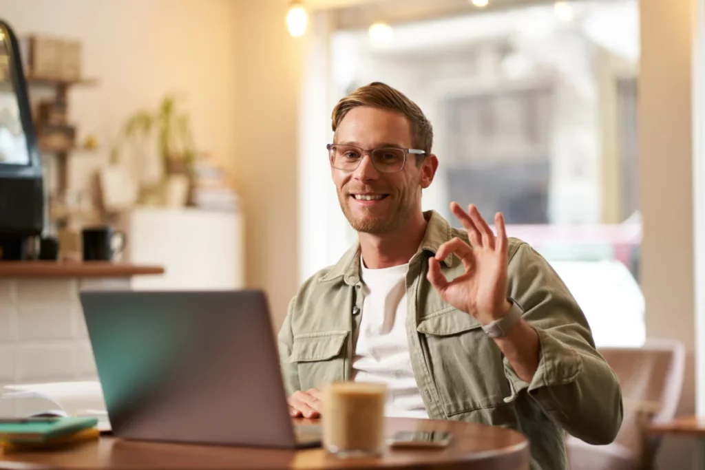 homem de negócios usando óculos e sorrindo ao mostra um sinal de ok, trabalhando no computador em um café - como fazer uma loja virtual