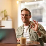 homem-de-negocios-usando-oculos-e-sorrindo-ao-mostra-um-sinal-de-ok-trabalhando-no-computador-em-um-cafe-como-fazer-uma-loja-virtual