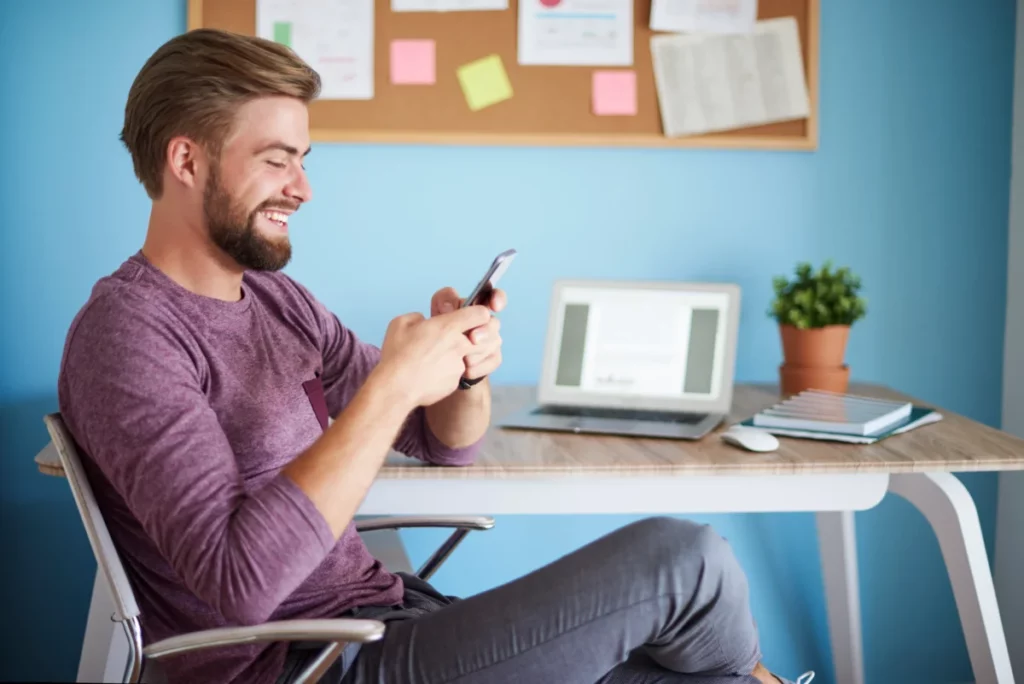 homem sentado mexendo no celular em frente ao computador em mesa de trabalho - atendimento via whatsapp