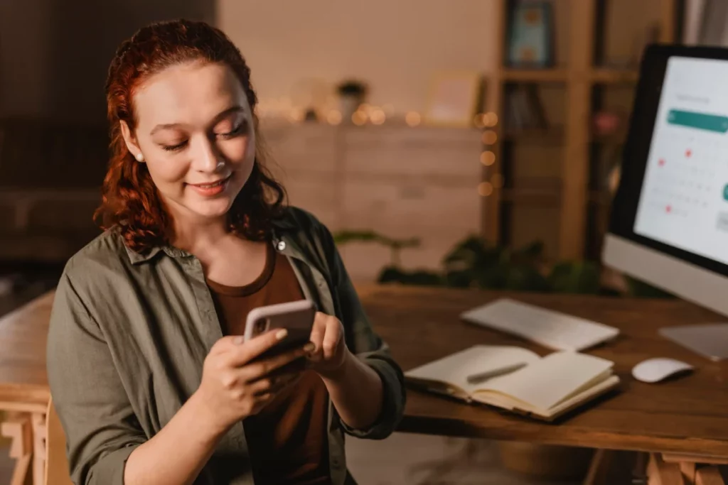 mulher sorrindo mexendo no celular em frente ao computador - atendimento via whatsapp