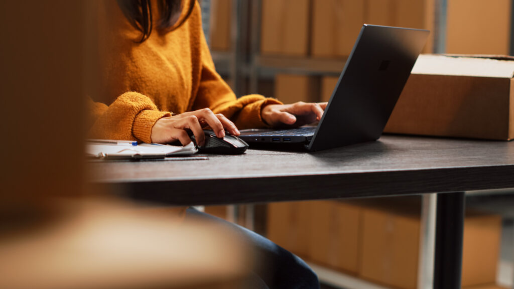Pessoa digitando em notebook em ambiente de estoque com caixas ao fundo, representando lojista gerenciando envios online