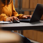 Pessoa digitando em notebook em ambiente de estoque com caixas ao fundo, representando lojista gerenciando envios online