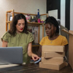 Duas mulheres analisando documentos ao lado de caixas de encomendas e um notebook, representando a gestão de envios de vendedoras online.