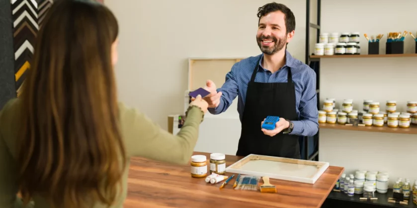 a-jovem-cliente-esta-entregando-ao-vendedor-sorridente-seu-cartao-de-credito-para-pagar-seus-suprimentos-de-arte-na-loja-pequenos-negocios-que-dao-certo-com-pouco-dinheiro