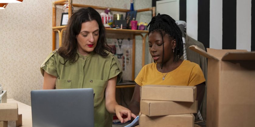 Duas mulheres analisando documentos ao lado de caixas de encomendas e um notebook, representando a gestão de envios de vendedoras online.